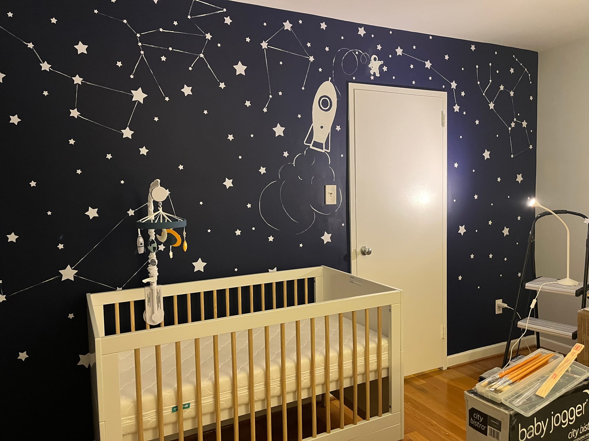 Space themed mural in the nursery. Dark blue background with hand painted constellations, a rocket ship, and astronaut in a spacesuit.